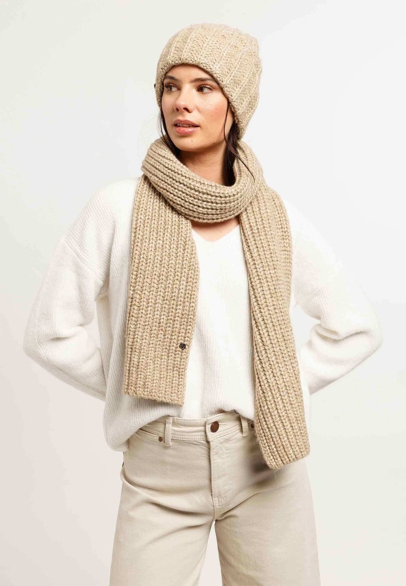 Set di berretto e sciarpa lavorati a maglia color beige, con una texture a costine. Indossato con un maglione bianco a maglia e pantaloni beige su uno sfondo chiaro.