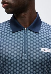 Κοντινό πλάνο ενός ατόμου που φορά μπλε polo με φερμουάρ, με σχέδιο, σκούρο γιακά και ετικέτα "Ted Baker Sport" στο στήθος.