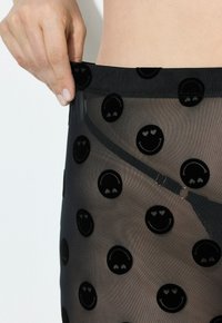Zwarte doorschijnende pantyhose met een patroon van verheven zwarte glimlachende gezichten. De elastische band is glad en benadrukt het speelse ontwerp.
