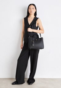 Femme portant un gilet sans manches à rayures fines noires et un pantalon large, tenant un sac à main noir structuré avec une bandoulière, debout contre un mur blanc.