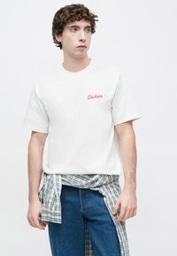 Λευκό βαμβακερό κοντομάνικο μπλουζάκι με κόκκινο λογότυπο "Dickies" στο στήθος. Φορεμένο με μπλε πουκάμισο καρό δεμένο γύρω από τη μέση.