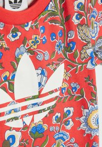 Havana košela s kvetinovým vzorom v červenej farbe, zdobená modrými a žltými kvetmi. Biele logo adidas a pruhy na prednej strane. Mäkko tkanina s okrúhlym výstrihom.