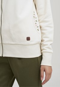 Sudadera cremosa con cremallera y bolsillo lateral, decorada con bordados florales en verde y rosa. Parche de logo marrón en el bolsillo. Textura de tela suave.