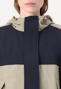 Veste à capuche bleu marine et beige clair avec une fermeture à boutons sur le devant et un logo brodé en bas à gauche. Tissu lisse avec un design en blocs de couleur.