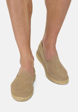 Gambe nude che indossano scarpe espadrillas slip-on color sabbia con suole intrecciate in juta su uno sfondo chiaro.