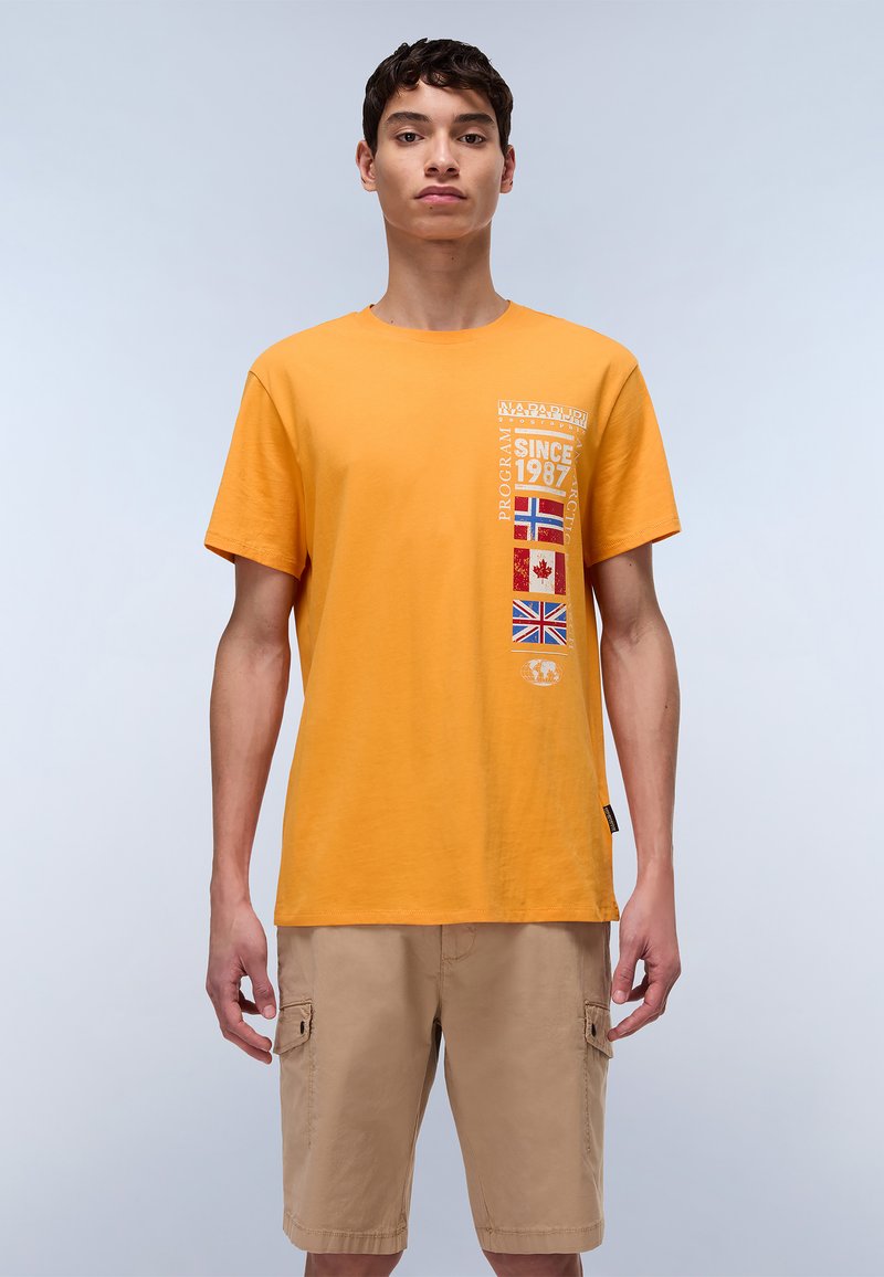 T-shirt en coton orange avec un design graphique représentant des drapeaux, le texte "SINCE 1987" et un col rond. Associé avec un short beige.