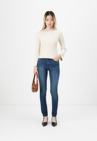Woman wearing a cream ribbed sweater, blue skinny jeans, black pointed heels, and holding a brown leather handbag, standing against a white background.