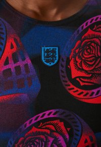 Svart t-skjorte med levende blomster- og geometriske mønstre i blått, rødt og lilla, med et blått skjoldlogo på brystet.