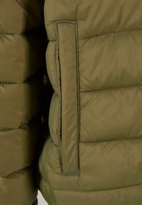 Vadderad jacka i olivgrön med en slät yta. Har en vertikal söm och en fickklaff på sidan för extra detalj.