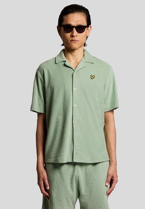 Mann trägt mintgrünes, strukturiertes, kurzärmeliges Hemd mit Knöpfen und passende Shorts, dazu dunkle Sonnenbrille, steht vor hellgrauem Hintergrund.