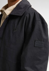 Jachetă neagră impermeabilă cu guler cu nasturi, textură subtilă și un logo brodat pe partea stângă a pieptului. Are un patch cu marcă pe mânecă.