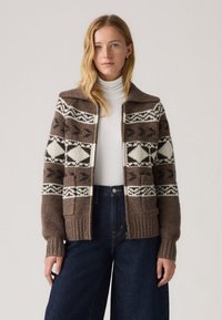 Rjav pleteni kardigan z geometrskimi belimi vzorci, z rebrastim ovratnikom in manšetami, podrobnostmi žepov, nošen čez belo polo majico, kombiniran s temnimi jeans.