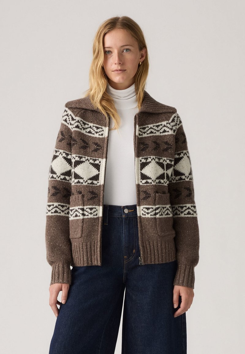 Brun strikket cardigan med geometriske hvide mønstre, ribbet krave og manchetter, lomme detaljer, båret over en hvid turtleneck, parret med mørk denim.