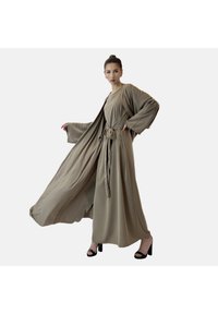 Robe beige longue et fluide avec de larges manches et un détail de taille noué, fabriquée en tissu lisse. Portée avec des sandales à talons noirs.