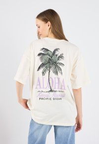 Camiseta color crema con un gráfico de palmera y "ALOHA" en morado. Con mangas cortas y un corte relajado, con texto impreso en la parte posterior.