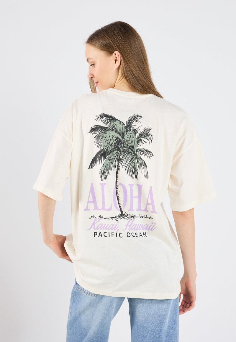 Camiseta color crema con un gráfico de palmera y "ALOHA" en morado. Con mangas cortas y un corte relajado, con texto impreso en la parte posterior.
