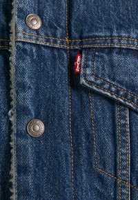 Giacca di jeans blu scuro, con cuciture arancioni e bottoni a pressione in metallo. Include un'etichetta rossa Levi's vicino alla tasca. Texture interna morbida.