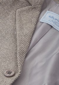 Primer plano de la solapa de una chaqueta de tela espiga en beige y gris, con un botón marrón y un forro interior liso de color gris claro con etiqueta cosida.