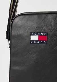 Čierna kožená taška na rameno s textúrovaným povrchom a výrazným nápisom "TOMMY JEANS" s červenými a bielymi akcentmi. Nastaviteľný popruh.