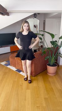 Fekete testhezálló felső és bővülő fekete szoknya. A modell sötét szandált visel, fapadlón áll, mögötte növény és texturált kanapé látható.