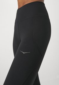 Hochgeschnittene schwarze Leggings aus dehnbarem Stoff, mit einem eleganten Design, einer glatten Oberfläche und einem kleinen reflektierenden Logo an der Seite.