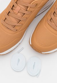 Braune Sportschuhe mit strukturiertem Obermaterial, Belüftungsöffnungen und weißen Akzenten. Zwei weiße Schnürsenkel, die unter den Schuhen positioniert sind.