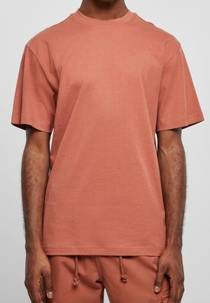Man die een bijpassend terracottakleurig T-shirt met korte mouwen en broek met trekkoord draagt, staand tegen een effen grijze achtergrond.