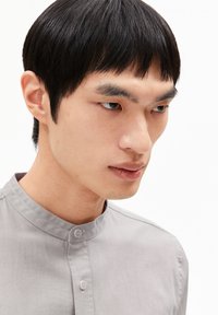 Grijze blouse met een textuurstof, voorzien van een knoopsluiting en een staande kraag. Het model heeft steil zwart haar en een neutrale gezichtsuitdrukking.