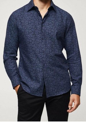 Homme portant une chemise à manches longues bleu foncé à motifs floraux avec un pantalon noir, debout avec une main dans la poche.