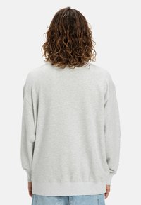 Persoon met schouderlang, krullend bruin haar, gekleed in een lichtgrijze sweatshirt en lichtblauwe spijkerbroek, met de rug naar de kijker op een witte achtergrond.