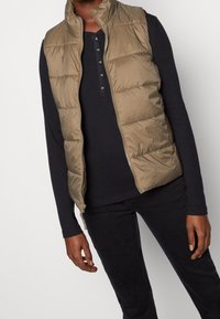 Beige Steppweste mit Reißverschluss, hohem Kragen und gestepptem Design, getragen über einem schwarzen Langarmshirt und dunklen Hosen.