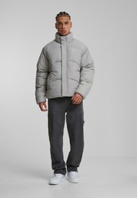 Chaqueta acolchada gris con alto cuello, diseño acolchado y cremallera frontal. El modelo lleva pantalones oscuros y zapatillas blancas. Branding sencillo visible.
