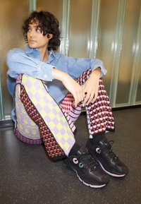 Mladý člověk s kudrnatými vlasy sedí na podlaze, má na sobě džínovou košili a barevné vzorované kalhoty a zamyšleně se dívá do strany.