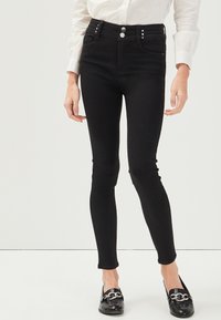 Jeans neri a vita alta con una trama liscia, caratterizzati da due bottoni frontali, borchie argentate sulla cintura e design slim-fit.
