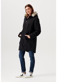 Černá parka bunda s odnímatelnou kapucí s kožíškem, na zip, s bočními kapsami, nošená přes modré džíny a bílé tenisky.