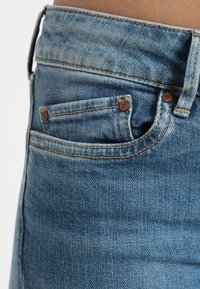 Los jeans de mezclilla azul claro presentan una cintura de altura media, diseño de cinco bolsillos, costuras en contraste y remaches metálicos en los bolsillos.