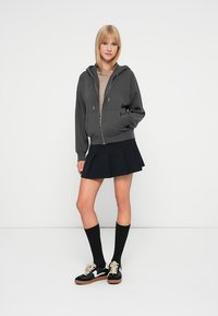 Tamno siva zip-up dukserica opuštenog kroja, nosi se preko bež majice. Uparena s crnom plisiranom suknjom i čarapama do koljena. Crne tenisice s bijelim detaljima.