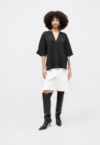 Sort oversized bluse med V-udskæring og korte ærmer, parret med hvide shorts og knæhøje sorte læderstøvler. Glat tekstur.