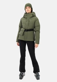 Groene gewatteerde jas met capuchon, rits- en knoopsluiting, riem en borstzak. Zwarte broek en skischoenen maken de look compleet.
