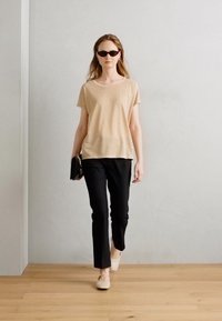 Beige T-shirt met korte mouwen, zwarte slim-fit pants, lichtgekleurde schoenen en een zonnebril, met een minimalistisch ontwerp tegen een neutrale achtergrond.