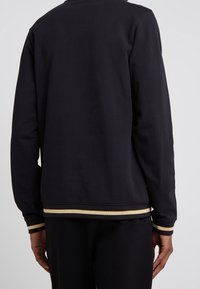 Sweatshirt noir avec des manches longues, des poignets côtelés et un ourlet à rayures dorées. Le tissu semble doux avec une finition légèrement texturée.