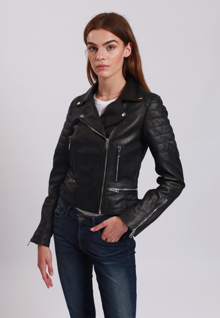 Giacca da motociclista in pelle nera con maniche trapuntate, cerniere argentate e tasche multiple. Indossata sopra una maglietta bianca e jeans blu.