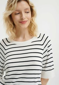 Witte gebreide top met zwarte horizontale strepen, met een ronde halslijn en korte mouwen. Zachte textuur, casual ontwerp en minimale details.