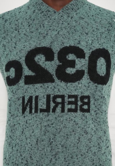 Groene gebreide vest met een gespikkelde textuur. Voorzien van oversized zwarte tekst met de opschrift "032C BERLIN" prominent op de voorkant.