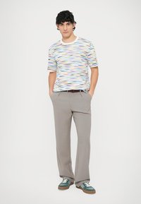 Kolorowy bawełniany t-shirt w faliste wzory, szare plisowane spodnie i zielone trampki. Model stoi z rękami w kieszeniach, neutralne tło.