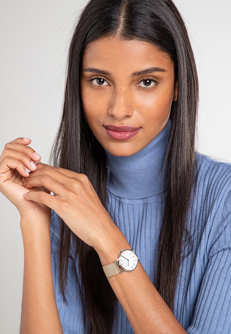 Zilveren mesh band horloge met een witte wijzerplaat en eenvoudige uurmarkeringen, gedragen om een pols tegen een achtergrond van een blauwe geribde trui.