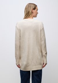 Maglione beige lavorato a maglia con maniche lunghe, polsini e orlo a costine, vestibilità rilassata e spacchi laterali, indossato sopra pantaloni blu scuro.