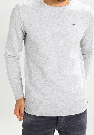 Mann trägt hellgrauen Pullover mit Rundhalsausschnitt und kleinem besticktem Logo auf der Brust und dunkelgraue Hose.