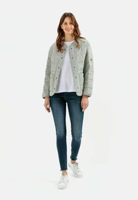 Hellgraue gesteppte Jacke mit Druckknöpfen, getragen über einem weißen T-Shirt, kombiniert mit dunkelblauen schmalen Jeans und weißen Sneakern.
