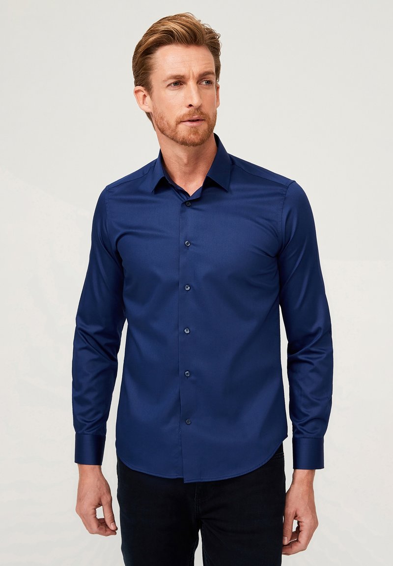 Marineblauwe blouse met lange mouwen, gladde textuur en een getailleerde pasvorm. Beschikt over een puntige kraag en bijpassende knopen.
